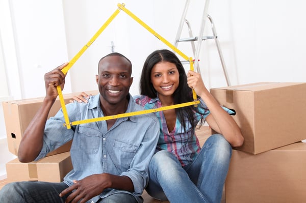 A man and a woman are sitting in front of boxes and holding a house-shaped frame around them