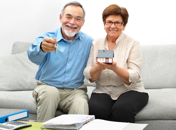 An older man and woman holding keys and a model house while sitting on a couch