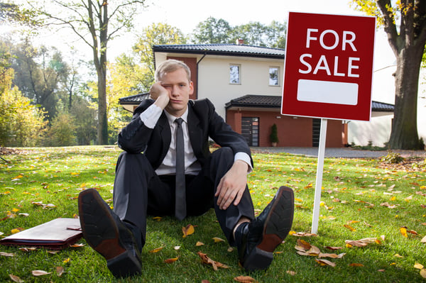 Real estate agent sitting dejectedly outside of a house with a for sale sign in the yard