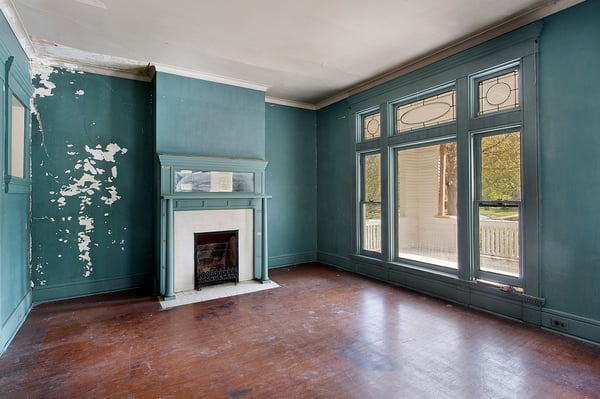 An empty room with a fireplace that has a damaged mantle, scuffed and dirty hardwood floors, and teal paint on the wall that has white showing through.