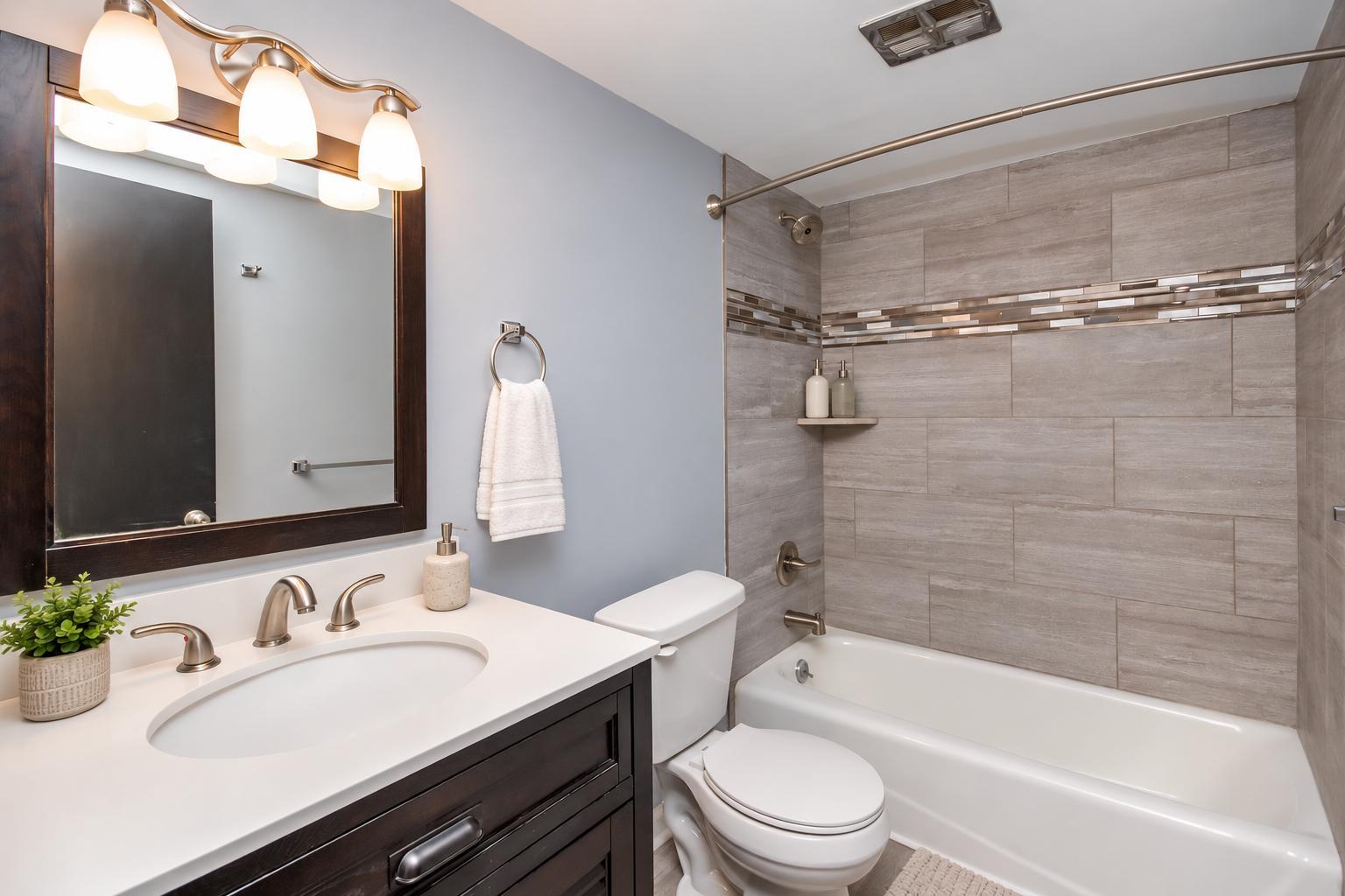 Renovated bathroom with dark wood vanity, framed mirror and tiled tub surround with a glass mosaic band